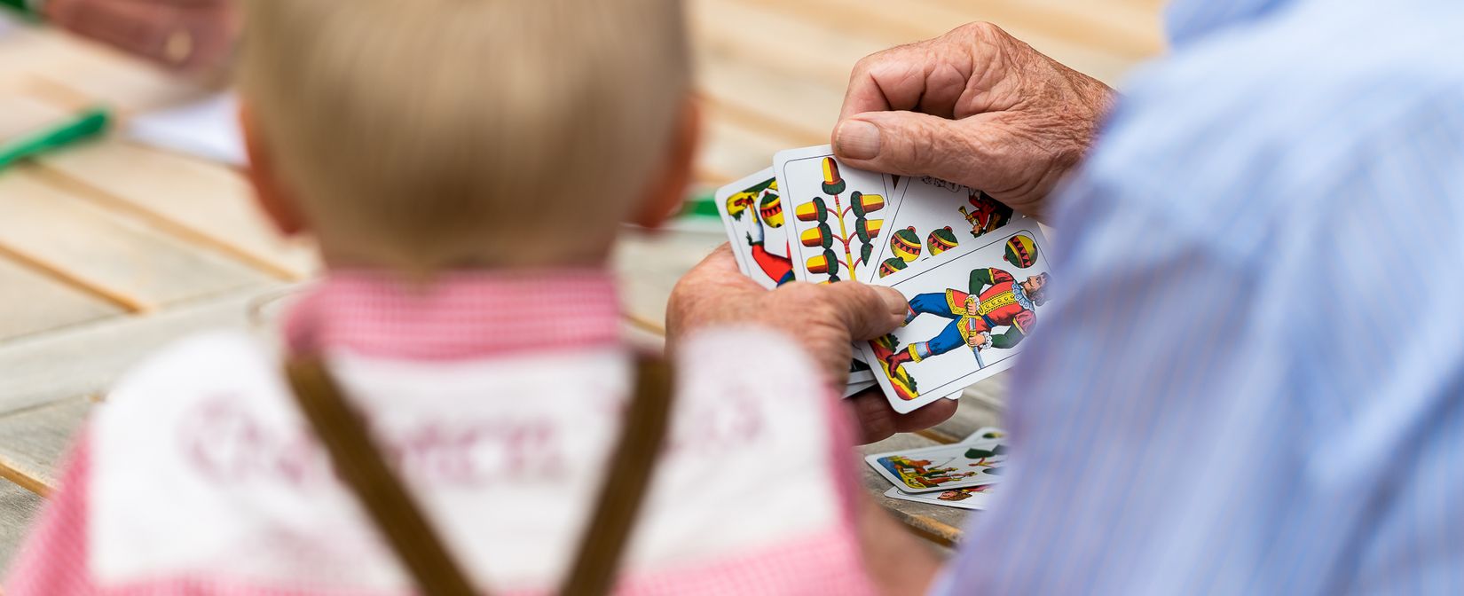 Generationen verbinden: Eine gemütliche Partie Schafkopf zwischen Opa und Enkel. Ein Kind in Trachtenkleidung und ein älterer Mann sitzen zusammen an einem Holztisch und spielen gemeinsam ein traditionelles Kartenspiel.
